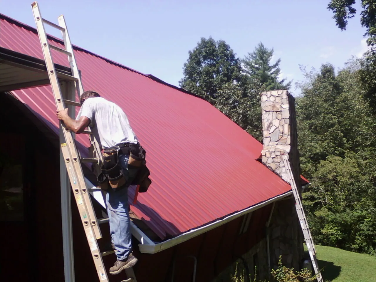 Roofer on a ladder beside a red metal roof and stone chimney — Soffit & Fascia Repair by Roofing Brothers in Dillon